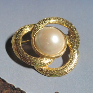 May include: A gold-toned brooch with a textured, intertwined design. The brooch features a large, round, cream-colored faux pearl at the center, set within a gold frame. The pin is a classic jewelry piece.