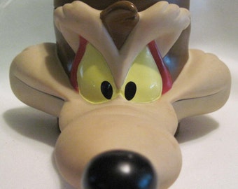 May include: A brown plastic cup shaped like Wile E. Coyote's head from the Looney Tunes cartoon. The cup has a black nose, yellow eyes, and a red mouth.