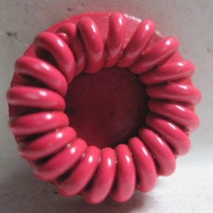May include: A bright pink plastic button with a raised, coiled design. The button has a smooth, circular center.