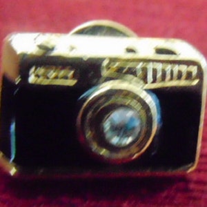 May include: A gold-tone camera pin with a black enamel body and a clear rhinestone lens.