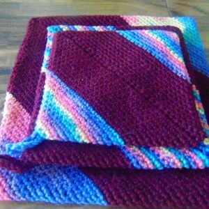 May include: Three crocheted pot holders in a stack. The top pot holder is a deep purple with a pink, blue, and white striped border. The middle pot holder is a light purple with a pink, blue, and white striped border. The bottom pot holder is a light blue with a pink, blue, and white striped border.