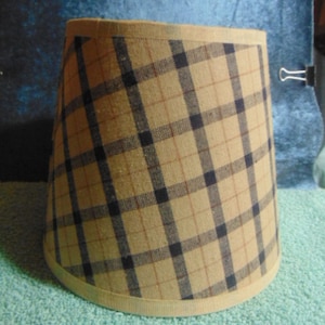 May include: A tapered drum lampshade featuring a tan fabric base with a dark navy blue and thin brown plaid pattern. The shade has a solid tan fabric trim along the top and bottom edges, creating a structured, cylindrical shape for a light fixture.