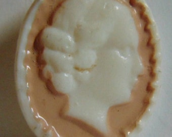 May include: A white cameo button with a raised profile of a woman's face. The cameo is set in a peach-colored oval frame with a scalloped edge.