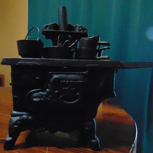 May include: A black cast iron toy stove with a small bucket and pots on top. The stove has a detailed design and sits on four legs. The stove has a handle on the side.