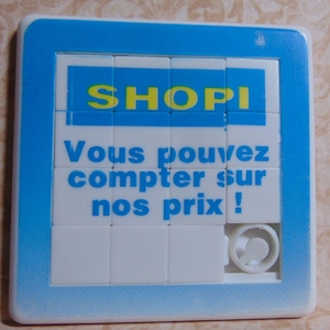 May include: A white plastic puzzle with a blue border. The puzzle has a picture of a blue sign with the word "SHOPI" in yellow letters. The sign also has the text "Vous pouvez compter sur nos prix!" in blue letters.