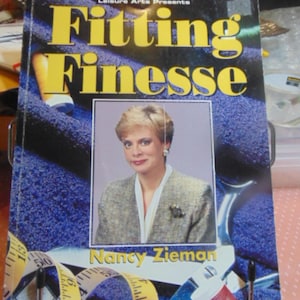 May include: A book titled "Fitting Finesse" by Nancy Zieman, presented by Leisure Arts. The cover features a woman in a blazer, set against a blue fabric background. A measuring tape and sewing notions are visible.