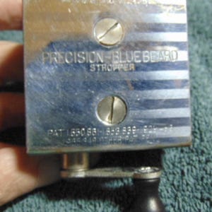 May include: A rectangular chrome-finished Precision-Bluebeard stropper with a hand crank and black knob. The metal surface features horizontal stripes, two flathead screws, and text reading MADE IN USA, PRECISION-BLUEBEARD STROPPER, and patent numbers.