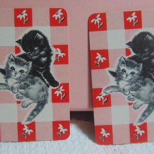 May include: Two rectangular cards with a red and white checkered pattern and white flower accents. Each card features an illustration of two kittens, one black and one gray, in a playful pose. The cards have rounded corners.