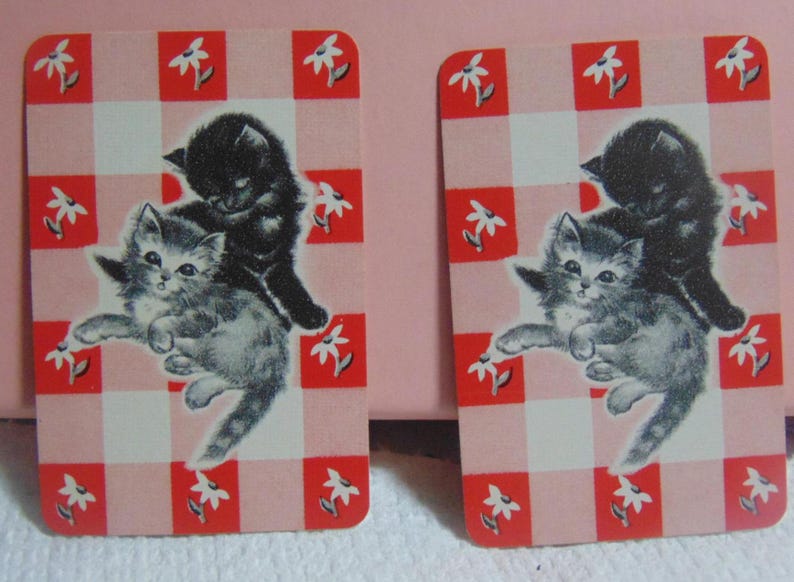 May include: Two rectangular cards with a red and white checkered pattern and white flower accents. Each card features an illustration of two kittens, one black and one gray, in a playful pose. The cards have rounded corners.