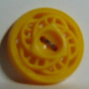 May include: A yellow plastic button with a decorative design. The button has a hole in the center for sewing.