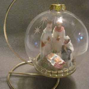 May include: A clear glass Christmas ornament with a gold stand. The ornament depicts the nativity scene with Mary, Joseph, and baby Jesus. The ornament is decorated with stars and a gold trim.