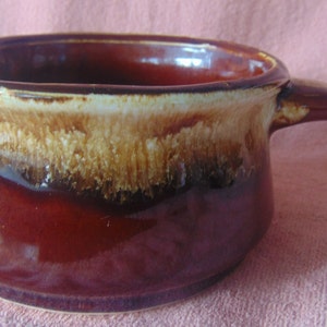 May include: A ceramic soup bowl with a handle. The bowl is a deep burgundy color with a gradient of tan and brown around the rim. The handle is curved and attached to the side of the bowl.