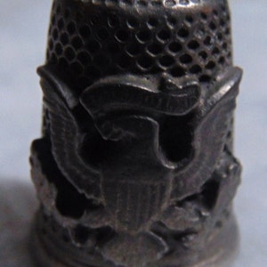 May include: A silver thimble with an eagle design. The thimble has a textured top and a smooth base.