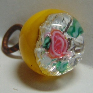 May include: A yellow glass button with a clear glass center featuring a pink rose and green leaves.