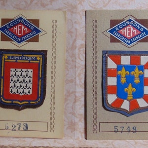 May include: Two vintage fabric patches with embroidered coats of arms. The first patch is brown with a red and blue shield featuring a white fleur-de-lis pattern. The second patch is blue with a red and white shield featuring three gold fleur-de-lis. Both patches have the text "ECUSSONS NEM FABRICATION FRANCAISE" and a number.