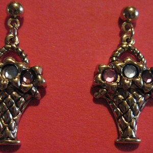 May include: A pair of gold-toned earrings featuring a basket design. Each earring has a basket shape with a handle and is adorned with three flower-shaped accents, each holding a gemstone in pink and clear colors.