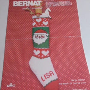 May include: A Bernat knitting pattern leaflet for a Santa's Heart stocking. The red cover features a white, green, and red knitted stocking with a Santa face, heart motifs, and the name LISA. Text includes Bernat, DMC, and size approx. 19 inches.