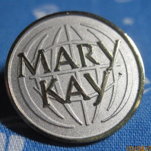 May include: A silver and gold round pin with the words "MARY KAY" and a stylized globe design.