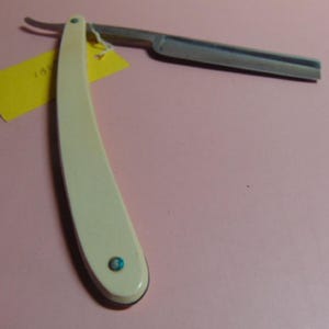 May include: An antique straight razor with a cream-colored handle and a silver blade. The handle has a small, round, blue-green accent near the end. A yellow tag is attached to the handle.