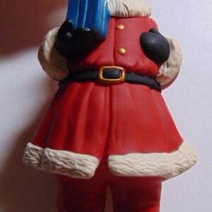 May include: A Santa Claus figurine wearing a red suit with a black belt and boots. The figurine holds a blue gift box. The suit has white fur trim. The Santa has a white beard and a hat.