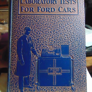 May include: A blue book with the title "Laboratory Tests for Ford Cars" embossed on the front cover. The cover features a blue silhouette of a mechanic working on a car engine.