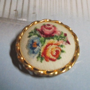 May include: A round, gold-toned brooch with a woven floral design. The center features a cream-colored fabric embroidered with a bouquet of roses in shades of pink, orange, and blue. The brooch has a twisted rope-like border.
