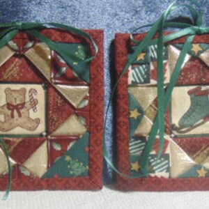 May include: Two square quilted fabric ornaments featuring a red, green, and gold geometric star pattern. One ornament displays a central image of a teddy bear with a candy cane, while the other shows an ice skate. Both have green ribbon ties.