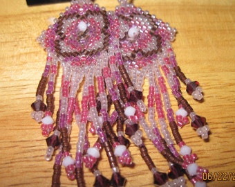 May include: A pair of dangle earrings with a beaded design. The earrings are made with brown, pink, and white beads. The earrings have a fringe of beads that hang down from the main design.