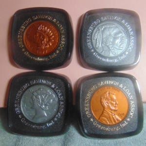 May include: Four square-shaped coin banks with different coin designs. Each bank has a dark gray border with the text "Greensburg Savings & Loan Assoc." and "Greensburg, Ind." The coins include an Indian head, a buffalo, and a penny.