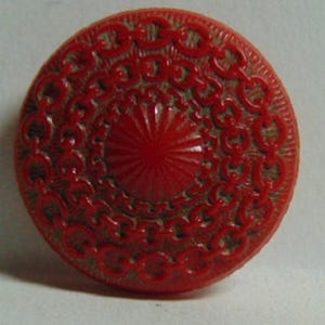 May include: A red plastic button with a detailed, intricate design. The button has a raised, circular center with radiating lines and a surrounding border of repeating patterns.