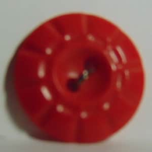 May include: A bright red button with a circular design. The button has a central hole and a pin, with white dots around the center. The button is a solid, vibrant red color.