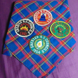 May include: A blue, red, and yellow plaid neckerchief with four circular patches. The patches feature text and images related to Greater N.Y. Councils Camporees from 1951, 1952, 1953, and 1954. The patches have a variety of colors, including green, red, and white.