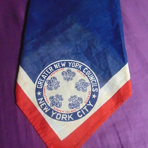 May include: A blue, white, and red vintage neckerchief. The neckerchief features a circular emblem with the text "Greater New York Councils" and the names of New York City boroughs. The neckerchief is folded in a triangle shape.