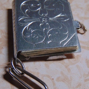 May include: A silver locket shaped like a book with an ornate floral design. The locket has a silver chain attached to it.