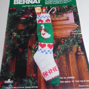 May include: A Bernat Holiday Goose knitted stocking pattern kit featuring a white stocking with a green band, red hearts, blue and white checkered accents, and a white goose graphic. The name ANNE is knitted in red on the toe. Size approx 19 inches.