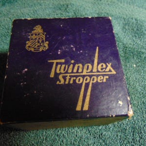 May include: A square, dark blue cardboard box featuring the gold-colored text Twinplex Stropper and a gold crest logo in the upper left corner. The box shows signs of wear along the edges and corners, with visible scuffing on the surface.
