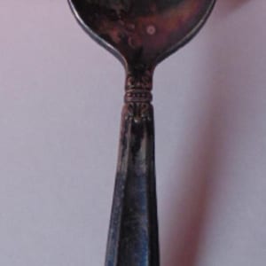 May include: Antique silver spoon with a rounded bowl and a decorative handle. The handle has a faceted design and ornate detailing at the base. The spoon is tarnished, showing its age and use.