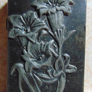 May include: A silver metal box with a floral design embossed on the front. The flowers are stylized and have a three-dimensional appearance.