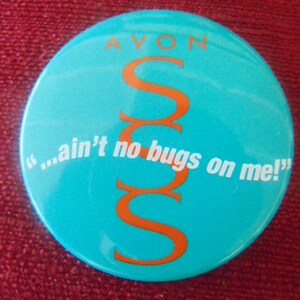 May include: A turquoise blue round button with the word "AVON" in white letters at the top. The letter "S" is repeated three times in orange, stacked vertically in the center of the button. The text "...ain't no bugs on me!" is written in white letters below the orange "S"s.
