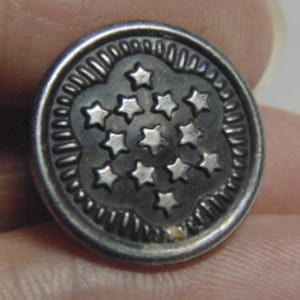May include: A round, antique silver button with a raised floral design in the center, containing eleven small, five-pointed stars. The button has a detailed, textured edge.