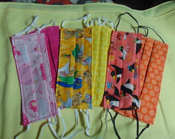 May include: Five fabric face masks with different patterns. The masks are pleated and have white elastic ear loops. The patterns include pink with white polka dots and a floral design, yellow with a blue sailboat and a cat design, yellow with white polka dots, pink with black penguins and a blue and red design, and orange with white polka dots.