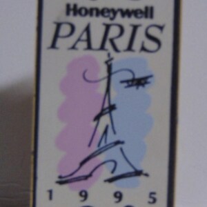 May include: A rectangular metal pin featuring a white background with the text Honeywell PARIS and 1995. The design includes a black line drawing of the Eiffel Tower with soft pink and light blue watercolor-style accents behind the tower.