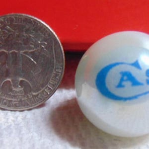 May include: A quarter coin next to a white marble with the word "CASE" in blue. The marble is approximately 1 inch in diameter. The coin is silver and has an image of a person on it.