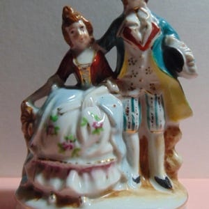 May include: A porcelain figurine depicting a couple in 18th-century attire. The woman wears a red and white dress with floral accents, while the man is in a blue coat and striped breeches. The base has a gold filigree design.