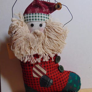 May include: A Christmas stocking ornament shaped like Santa Claus. The red stocking has a black and red checkered pattern, green buttons, and a green heel. Santa has a red hat with white snowflakes, a white beard, and a button on the hat.