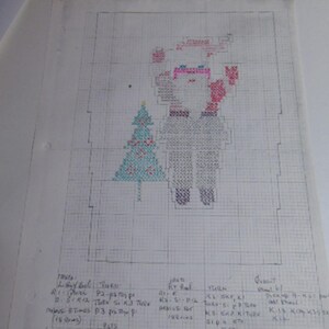 May include: A white sheet of graph paper featuring a hand-drawn cross-stitch pattern of a Santa Claus figure next to a Christmas tree. The design uses red, blue, and black ink. Below the graphic, handwritten knitting instructions are clearly visible.