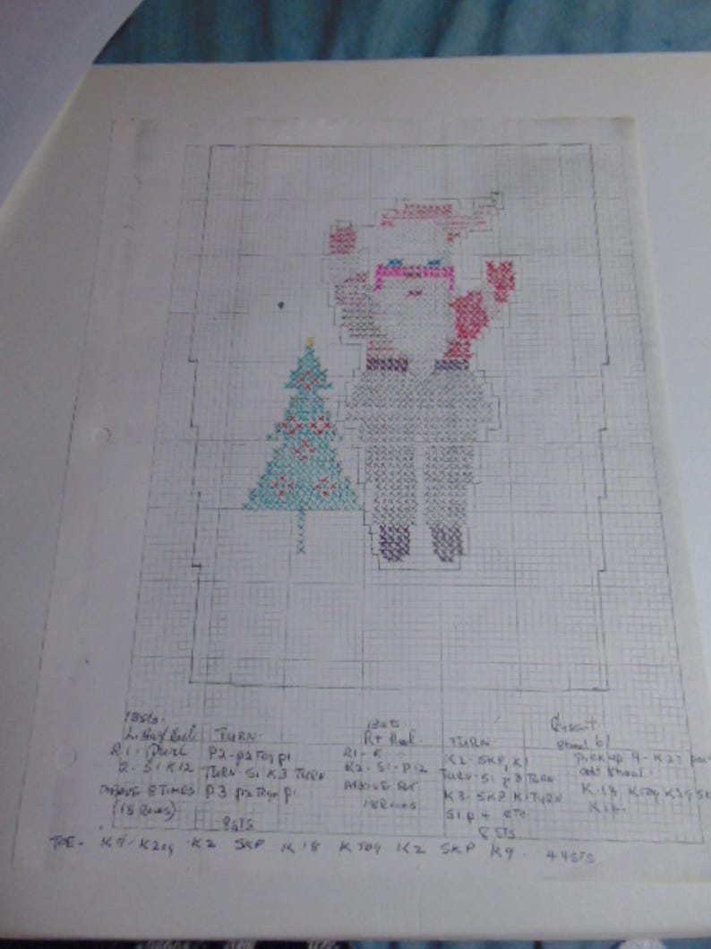 May include: A white sheet of graph paper featuring a hand-drawn cross-stitch pattern of a Santa Claus figure next to a Christmas tree. The design uses red, blue, and black ink. Below the graphic, handwritten knitting instructions are clearly visible.