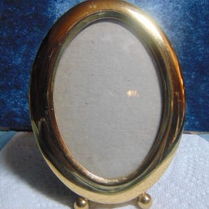 May include: An oval-shaped picture frame with a polished gold-tone metal finish. The frame features a smooth, rounded border and is supported by two small, spherical metal feet at the base, allowing it to stand upright on a flat surface.