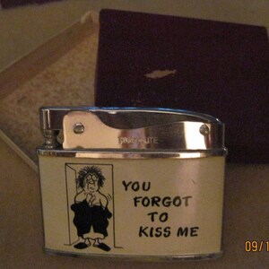 May include: A white lighter with a silver top and a cartoon illustration of a person peeking out of a doorway. The lighter has the text "You forgot to kiss me" printed on it.