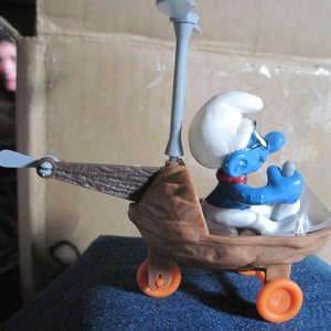 May include: A blue Smurf figurine is piloting a brown toy helicopter with a propeller on top. The helicopter has two wheels and a brown wooden-like body.